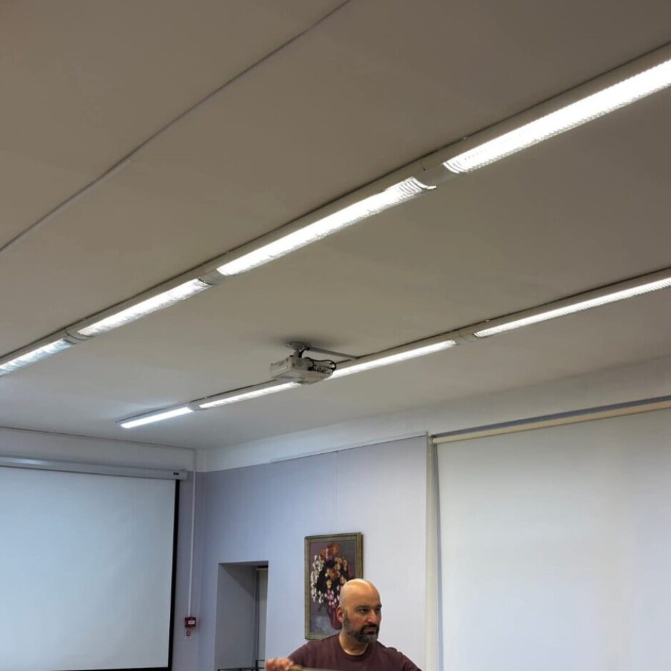 Vīrietis ar diriģenta zizli rokā vada orķestri vai kori mēģinājumu telpā ar baltām klavierēm un projektoru ekrāniem.